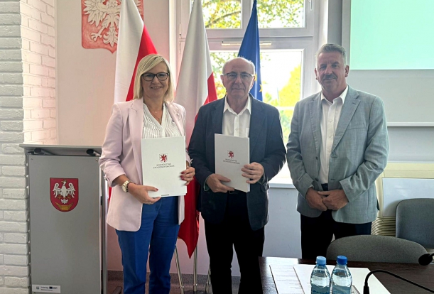 W Mirosławcu powstanie rodzinny dom dziecka. Strony porozumienia prezentuja podpisany dokument, w tle okna, flagi i telebim.