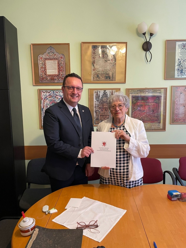 Bogdan Jaroszewicz i Róza Król trzymaja teczkę z podpisaną umową. W tle wiszą stare zdjęcia na żółto-zielonej ścianie