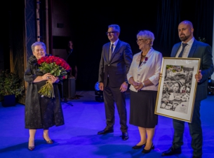 Małgorzata Usielska z naręczem kwiatów stoi na scenie, obok trzy osoby, jedna z nich trzyma dużą grafikę w ramie