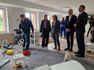 goście spotkania stoją w sali i rozmawiają, na dywanie siedzą dzieci z opiekunami