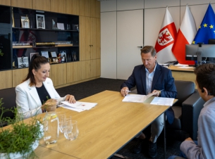 Strony porozumienia podpisują umowę przy stole prezydialnym, w tle, ściany i gabloty. 