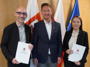 Marszałek Geblewicz wraz beneficjentami prezentują podpisaną umowę, w tle drewniana ściana i flagi.