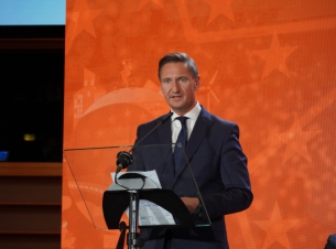 marszałek województwa Olgierd Geblewicz stoi przy mównicy i przemawia, w tle pomarańczowe tło