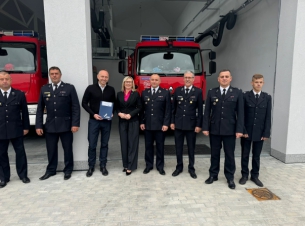 grupa druhów OSP Dygowo wraz z Anną Bańkowską i wójtem Grzegorzem Starczykiem stoja przed remizą. W tle dwa wozy strażackie