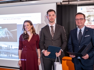 Członek Zarządu Województwa Bogdan Jaroszewicz pozuje do zdjęcia z laureatem i członkinia jury, w tle wizualziacja pracy laureata i plakat konkursu. 