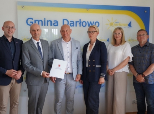 Strony porozumienia prezentują podpisany dokument, w tle ścianka promocyjna Gminy Darłowo. 