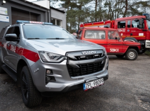 srebrny samochód terenowy z napisem straż, w tle dwa czerwone wozy strażackie; pojazdy stpja przed remizą 