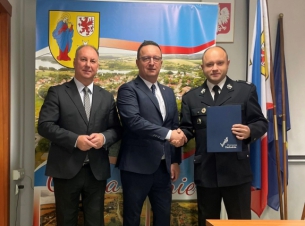 wspólne zdjęcie wójtak, członka zarządu i druha OSP na tle kolorowej ścianki promocyjnej, druh trzyma w jednej dłoni umowę, druga podaje członkowi zarządu województwa 