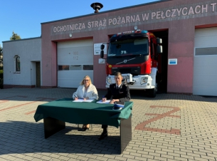 przed remizą strażacką stoi stolik; przy stoliku siedzi druh osp Pełczyce i Anna Bańkowska, podpisują umowę