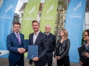 Marszałek Geblewicz składa podpis na umowie, w tle budynki, inne osoby uczestniczące w konferencji.