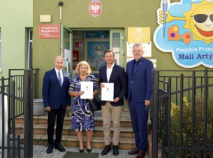 Sygnatariusze umowy prezentuja podpisany dokument, w tle budynek przedszkola. 