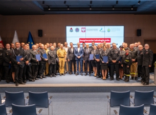 Strażacy z umowami i gośćmi konferencji stoją na scenie i prezentują podpisane dokumenty, w tle ekran z tytułem konferencji.