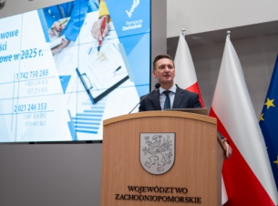 Marszałek Geblewicz przy mównicy prezentuje założenia budżetu na 2025 rok, w tle telebim ze slajdem o budżecie. 