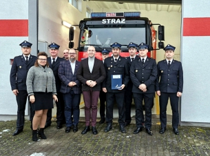 Wicemarszałek Jakub Kowalik wraz ze strażakami-ochotnikami pokazują do wspólnego zdjecia przed remizą, w tle wóz strażacki.