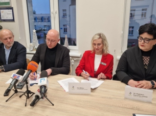 Strony umowy podpisuja dokument za stołem preydialnym, w tle okna i widok na budynki szpitala w Szczeicnku. 