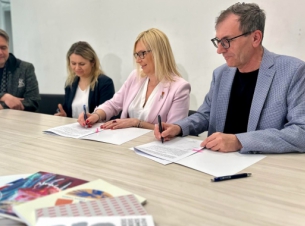 Sygnatariusze umowy podpisują dokument przy stole prezydialnym, w tle ściana i ekran ze zdjęciem.