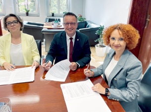 Strony porozumienia podpisują przy stole dokument, w tle ściana i okna pomieszcenia.