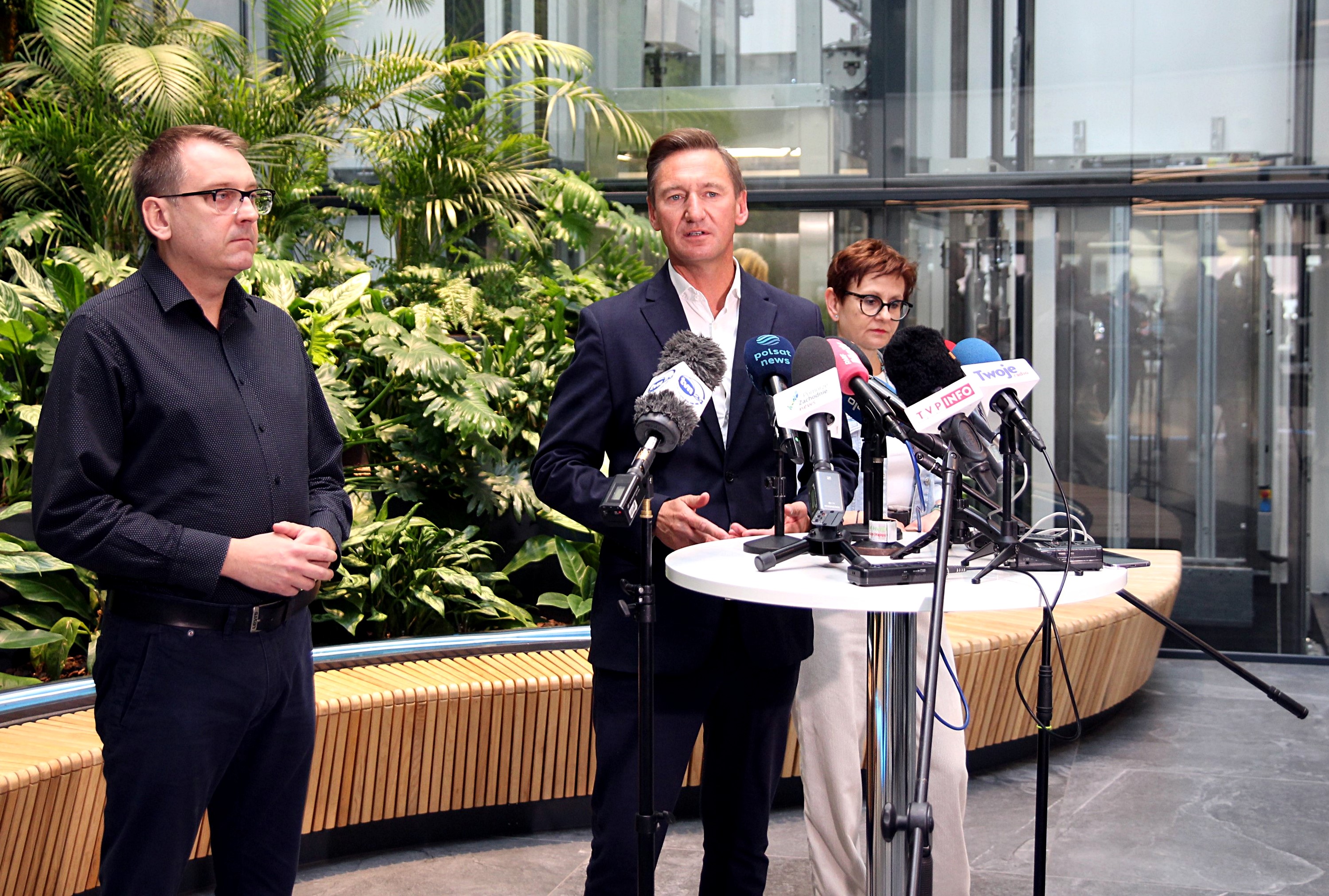 Dawid Krystek, Olgierd Geblewicz i Gabriela Wiatr podczas konferencji prasowej. Stoją na tle roslinnej ściany i niebieskiego panelu z wodą